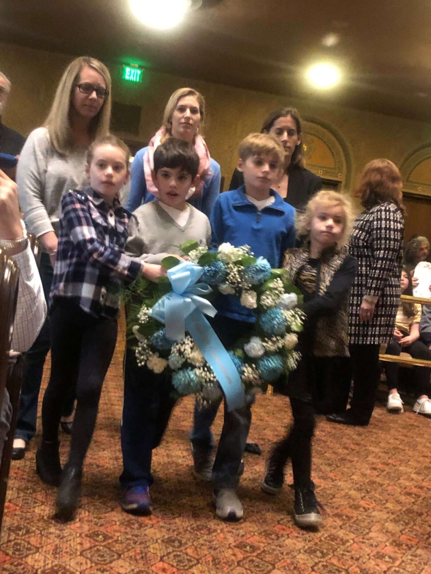Children holding floral wreaths at a memorial event indoors.